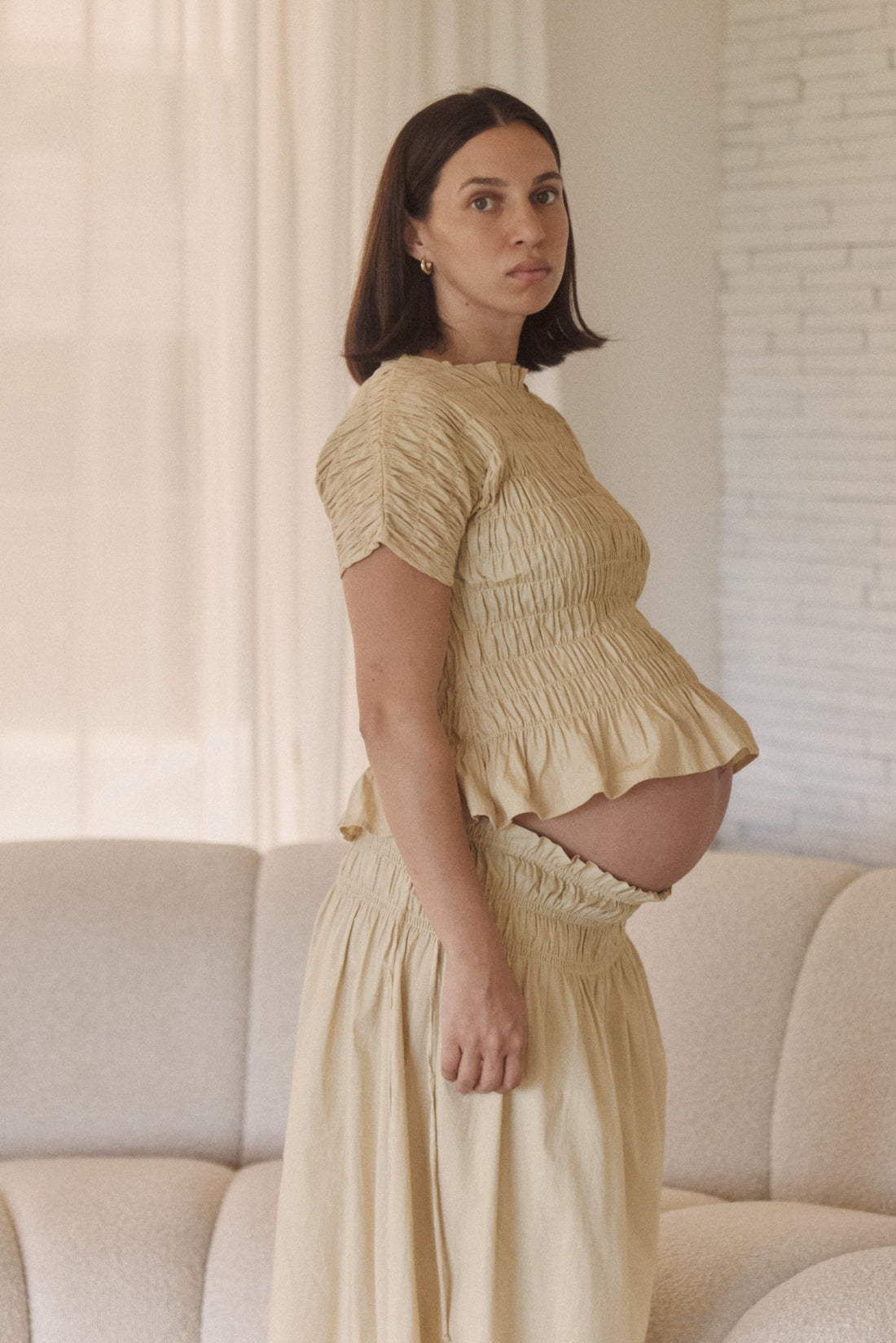 Pregnant woman in a light yellow outfit standing in a room with a white couch and light-colored walls.